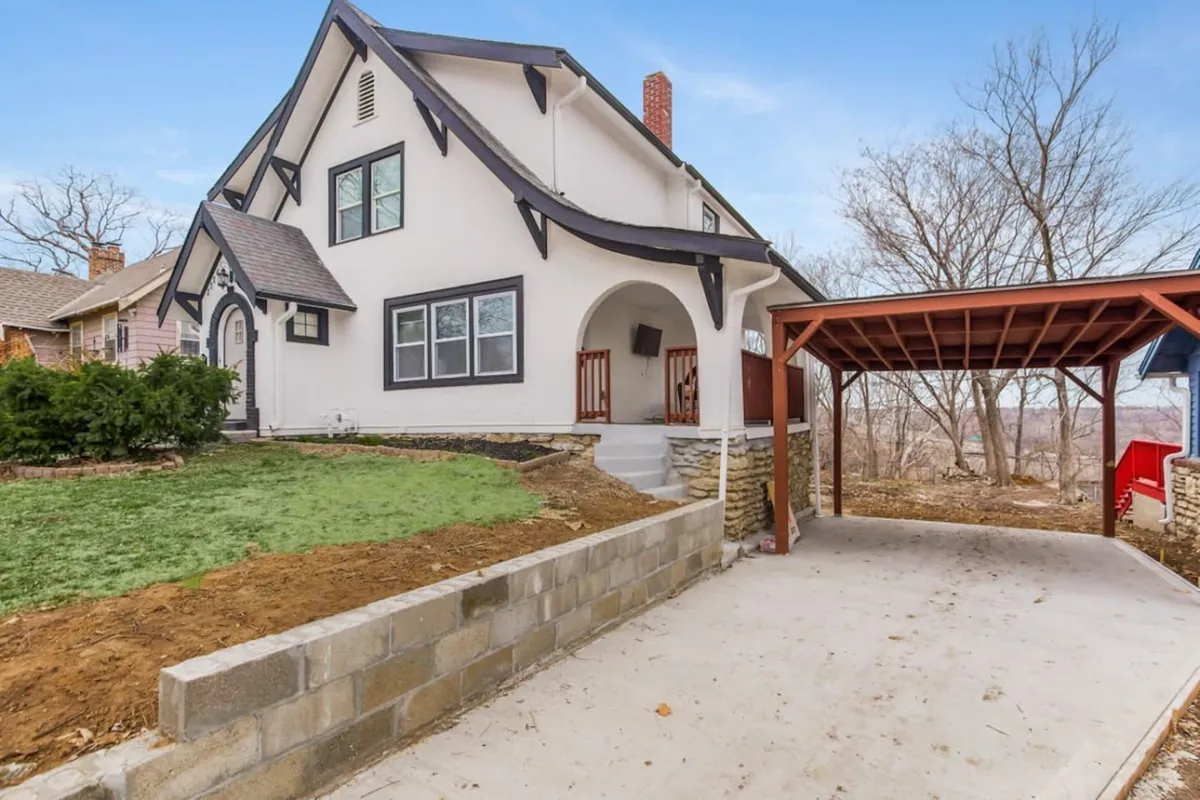 The hero image features the exterior of a spacious single-family house in Kansas City, Missouri, showcasing its unique architectural design, detailed stonework on the foundation, and a practical carport. The inviting porch is perfect for lounging, and the lush yard offers ample space for outdoor entertaining and gatherings.
