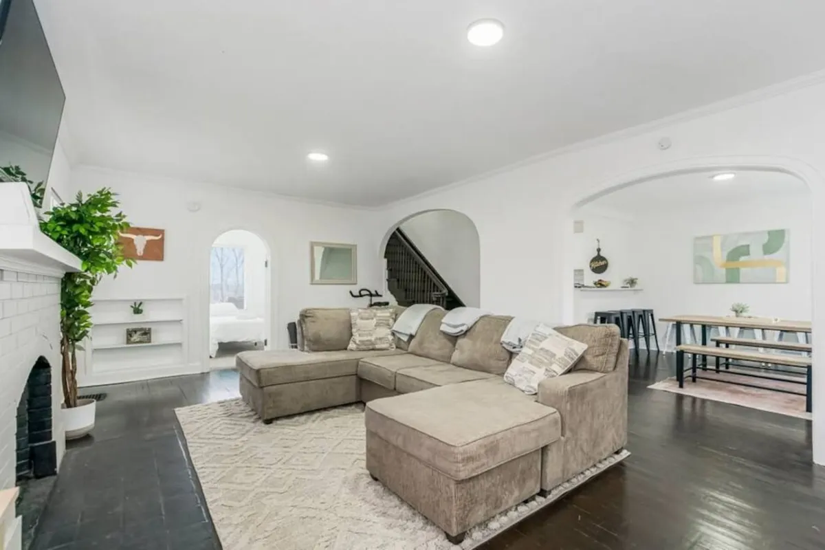 This image features a large sectional sofa positioned near a modern fireplace, accented by a decorative plant and artwork on the wall. A staircase and multiple seating options suggest an open, inviting space conducive to group gatherings or casual collaboration. The overall mood is comfortable and welcoming, making it ideal for team relaxation or informal meetings after a day of business activities.