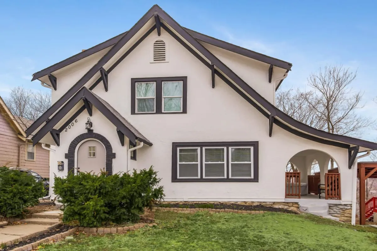 The front driveway of the home, framed by manicured shrubs, a unique roofline, large front windows, and an inviting arched entryway. The spacious driveway is visible, clearly capable of fitting multiple vehicles for group arrivals and departures.