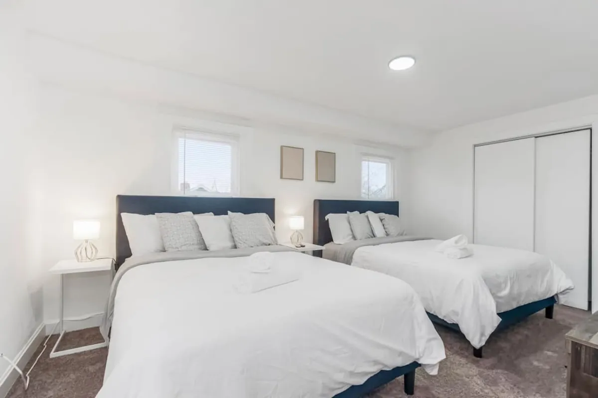 This serene bedroom scene displays two beds with blue headboards, small bedside tables with lamps, and minimalist wall art. Natural light filters into the room, creating a peaceful, restful environment. The space feels both practical for groups and intimate for private relaxation.