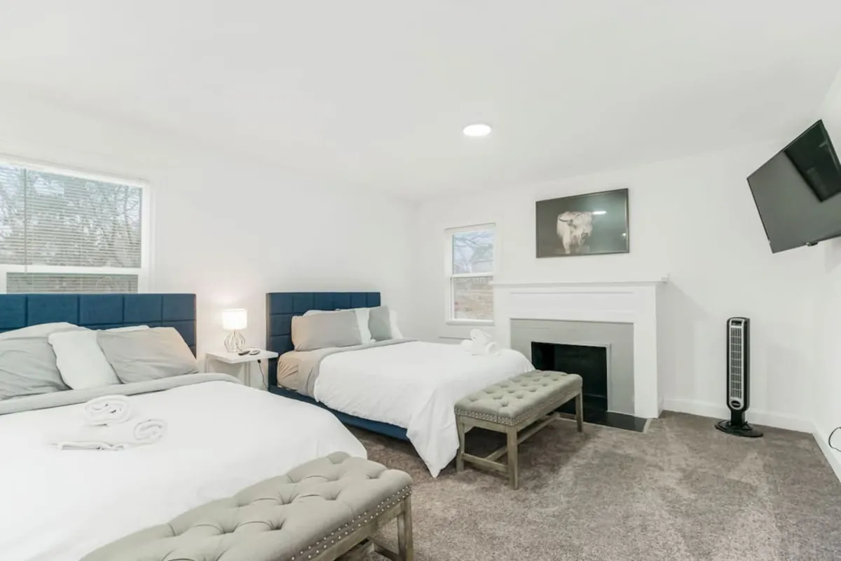 This image features two beds with blue headboards set in a spacious, well-lit bedroom. There's a fireplace that adds a cozy touch, a wall-mounted TV positioned for easy viewing from bed, and a bench at the foot of each bed. The overall mood is comfortable and inviting, designed for rest and relaxation.