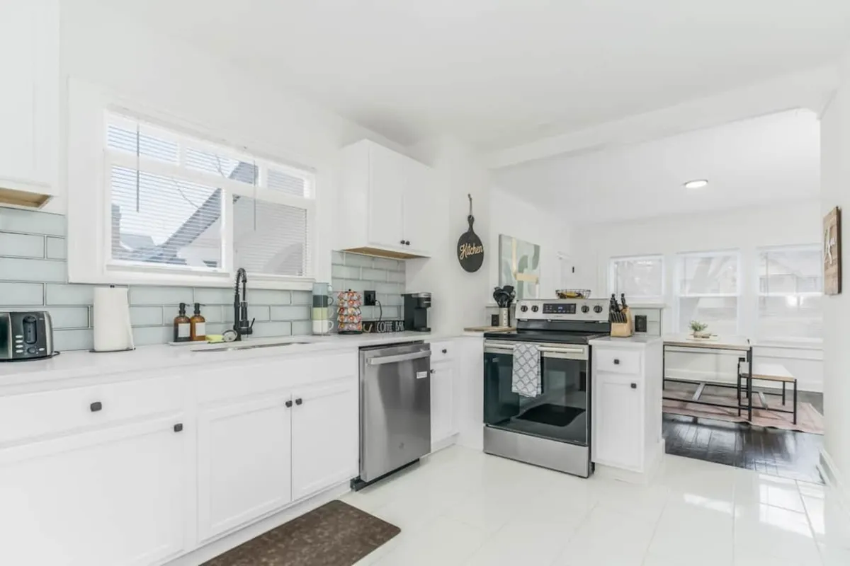 A bright, modern kitchen featuring sleek white cabinetry, a spacious kitchen island perfect for group food prep, modern appliances, decorative wall art, and window views that lend natural light to the space. The design reflects both functionality and style, ideal for entertaining and communal cooking.