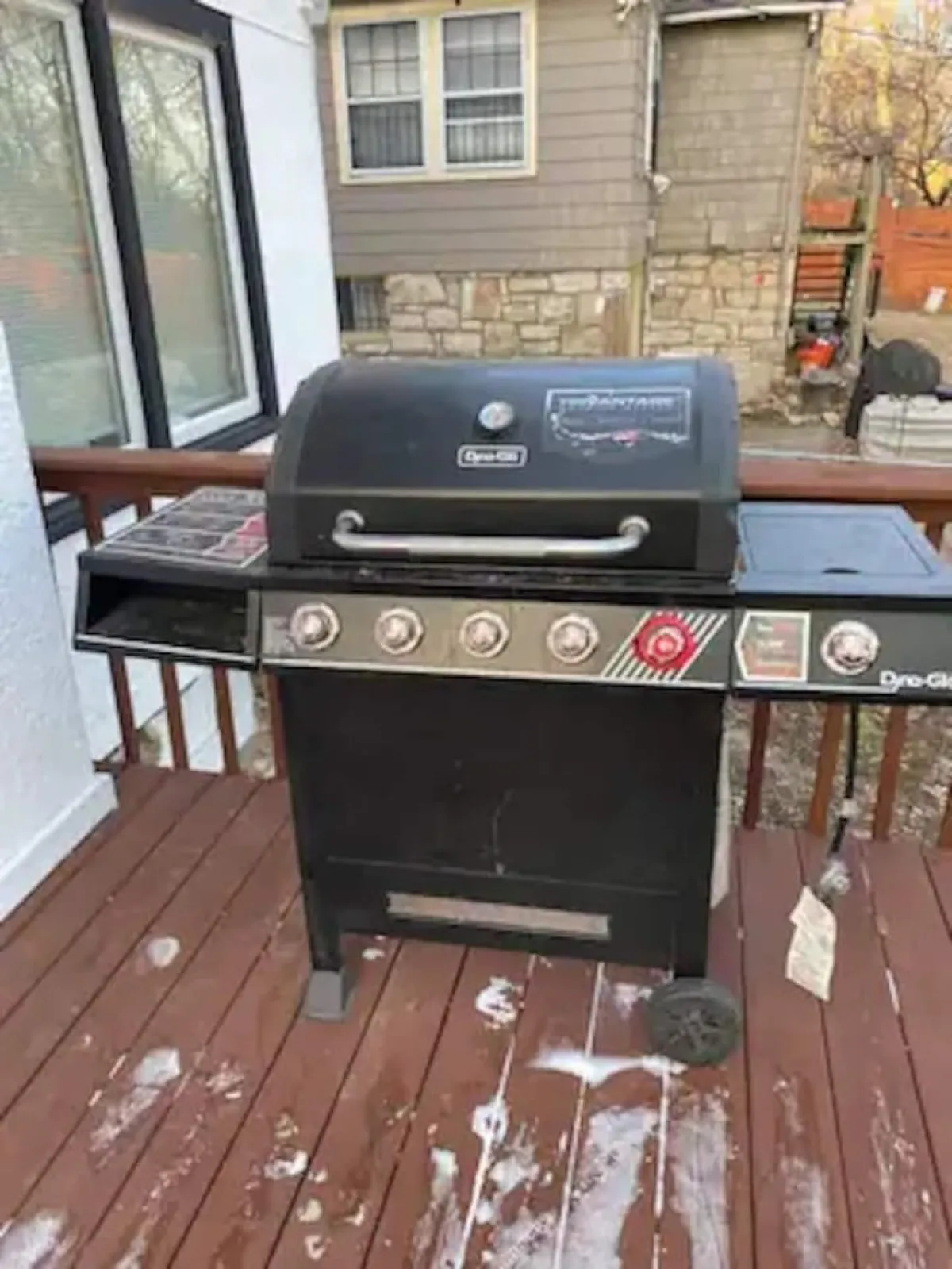An outdoor deck area featuring a grill, seating, and a view of the surrounding house. The space is clearly designed for outdoor dining, barbecuing, and social gatherings, suggesting a smooth transition from kitchen to patio for al fresco meals.